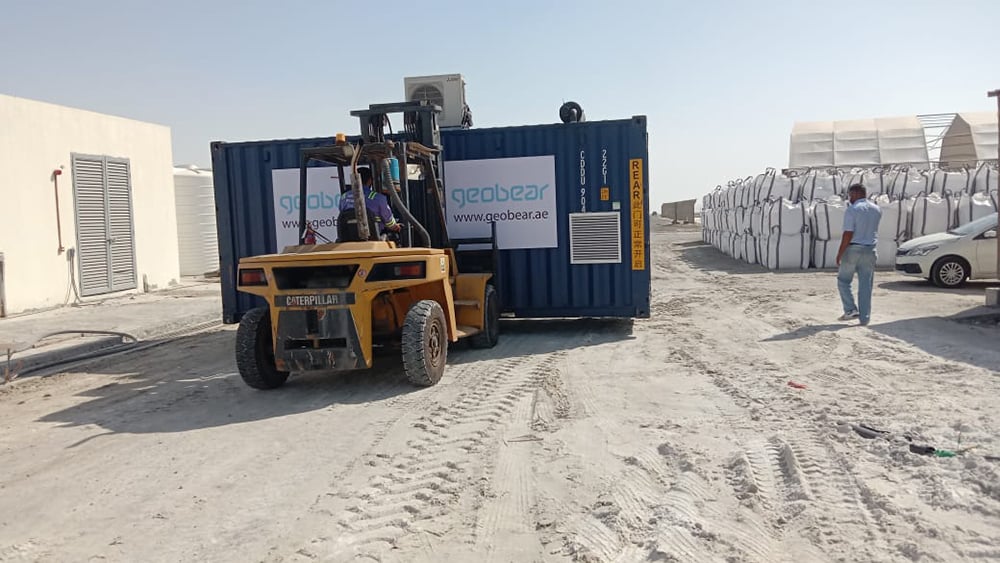 Geobear treatment container being carried to site via front loader on sandy ground.