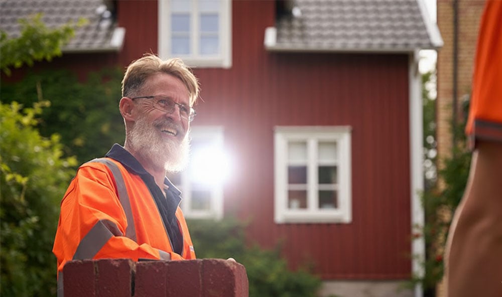 Geobears tekniker i orangea kläder ler framför ett rött trähus med vita knutar.