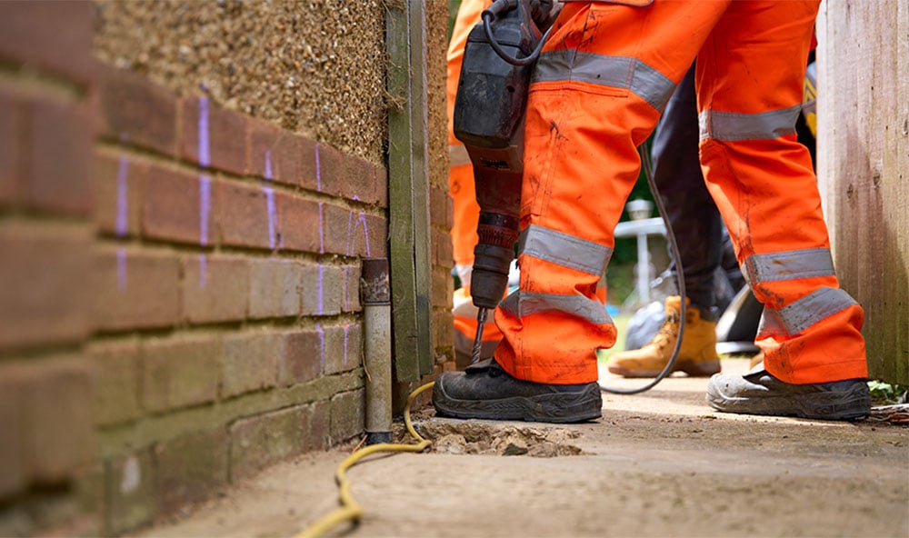 Benen på Geobears tekniker i orangea kläder håller i borr vid en husvägg/tegelvägg.