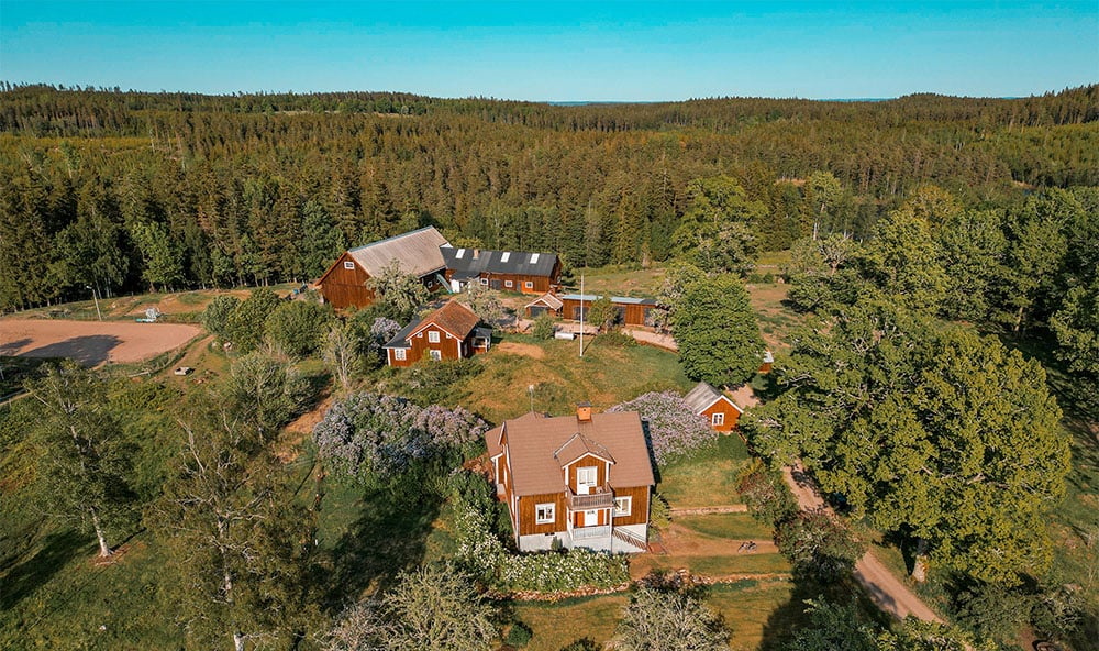 Drönarbild över ett svenskt lantligt område med mycket skog, röda hus och soligt väder.
