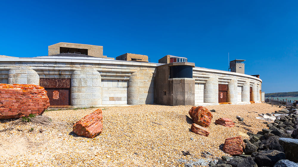 Exterior view from ground level of a section of Hurst castle with large red rocks and pebble cover in foreground.