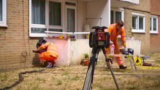 View looking past a rotating laser level on a tripod to Geobear techs working on raising a sunken terrace in a block of flats.