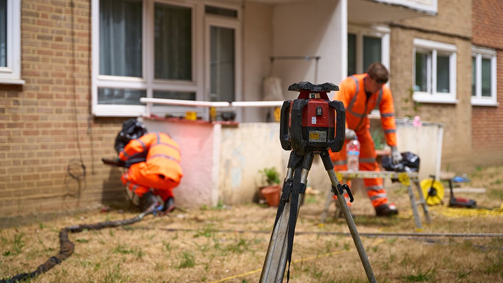View looking past a rotating laser level on a tripod to Geobear techs working on raising a sunken terrace in a block of flats.