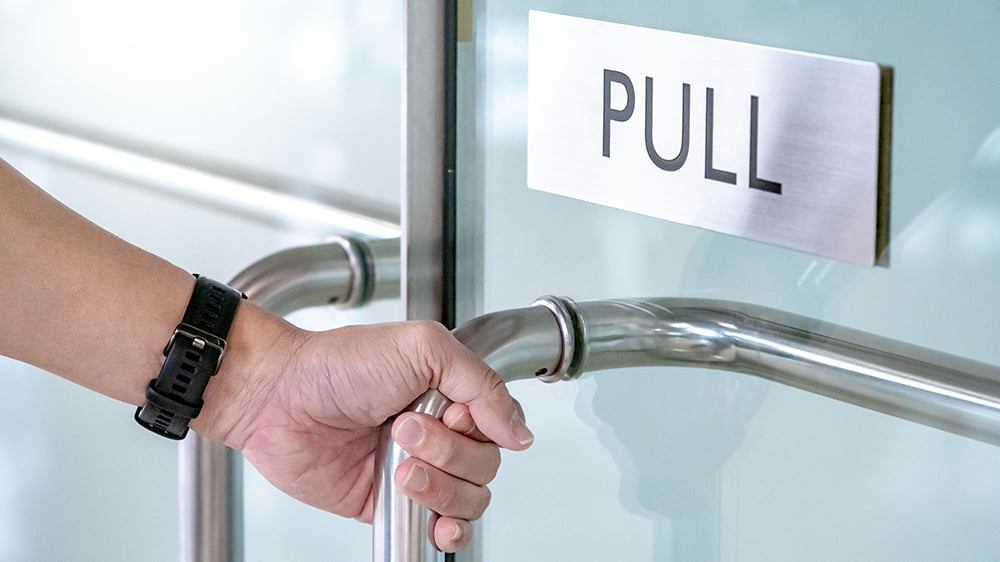 Hand holding on a metal handle on a glass door with the sign reading "pull"